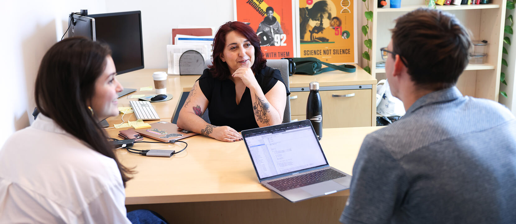 A woman sits behind a desk talking with two younger employees